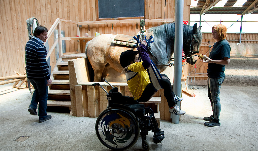 Aufsatteln per Knopfdruck: Auf Hof Grüneberg hilft ein Lift Menschen mit Mobilitätscheinschränkungen aufs Pferd, Foto: Studio Prokopy Ein Sepziallift hebt ein Kind aus dem Rollstuhl und aufs Pferd.