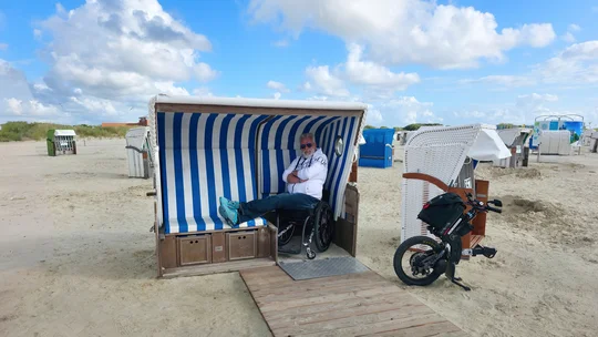Reiseblogger Hans-Jürgen rollt im barrierefreundlichen Strandkorb in Ostfriesland Reiseblogger Hans-Jürgen sitzt mit ausgestreckten Beinen im Strandkorb, daneben steht sein Zuggerät