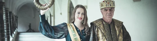 eine junge Frau hält einen goldenen Kranz in die Höhe. Sie trägt eine Scherpe. Neben ihr ein älterer Herr mit Krone