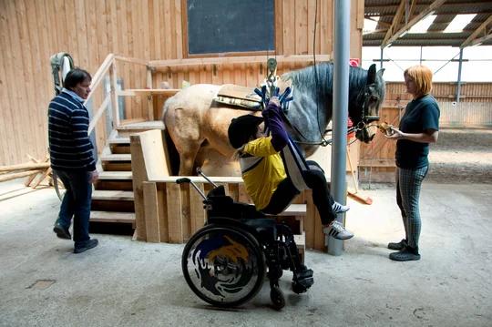 Aufsatteln per Knopfdruck: Auf Hof Grüneberg hilft ein Lift Menschen mit Mobilitätscheinschränkungen aufs Pferd, Foto: Studio Prokopy Ein Sepziallift hebt ein Kind aus dem Rollstuhl und aufs Pferd.