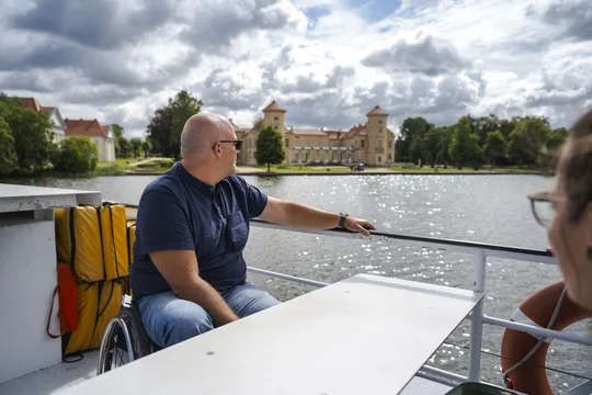 Aussicht an Deck © Madlen Krippendorf Ein Rollstuhlfahrer sitzt auf dem Deck eines Ausflugsschiffs und guckt auf das Schloss Rheinsberg.