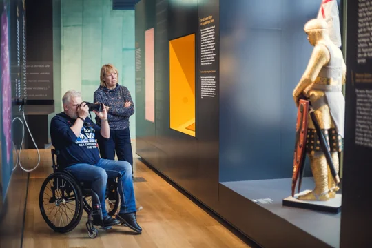 Rollstuhlfahrer fotografiert eine Ritterstatue die in einem modern beleuchteten Gang im Dommuseum Ottonianum steht. Neben ihm steht eine interessierte Frau und sieht sich ebenfalls die Statue an.