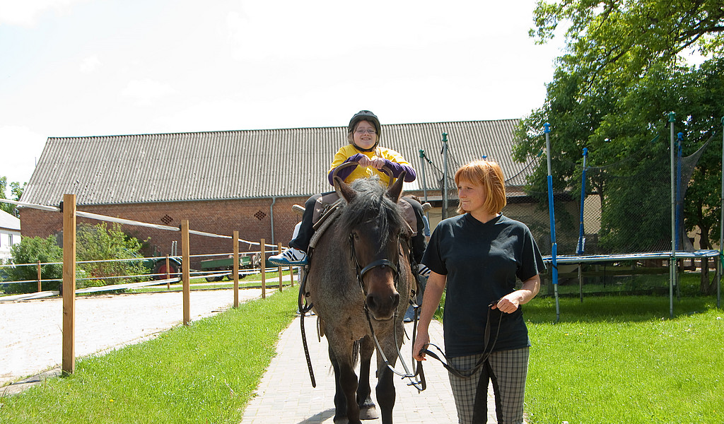 Ausritt auf Hof Grüneberg, Foto: Studio Prokopy Ein Mädchen reitet auf einem Pferd, dass von der Reitlehrerin geführt wird. Links und rechts ist Pferdekoppel, im Hintergrund der Stall.